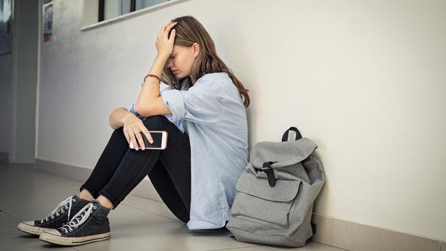 Upset and depressed girl holding smartphone sitting on college campus floor holding head. University sad student suffering from depression sitting on floor at high school. Lonely bullied teen in difficulty with copy space.