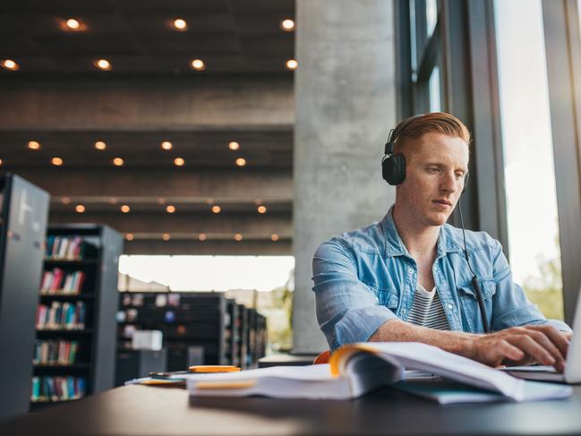 Young male student with headphones studying on the laptop. Handsome caucasian man sitting at the desk and working on laptop at public library. Picture: Istock