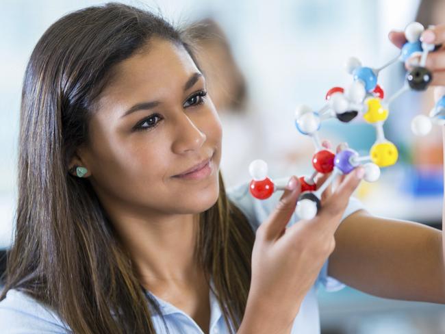 Teenage Hispanic female high school student is using plastic educational model toy molecules while studying in private school science class. Girl is holding study material while talking to teacher. Student is wearing a private school uniform.