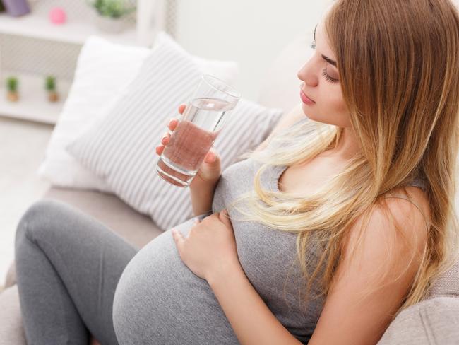 Pregnant woman with glass of water sitting on sofa closeup. Young expectant blonde rest at home. Pregnancy, healthcare, thirst concept Picture: Istock