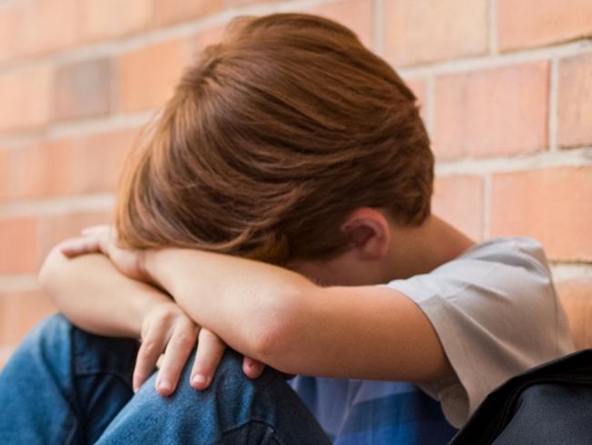 Little boy sitting alone on floor after suffering an act of bullying while children run in the background. Sad young schoolboy sitting on corridor with hands on knees and head between his legs.