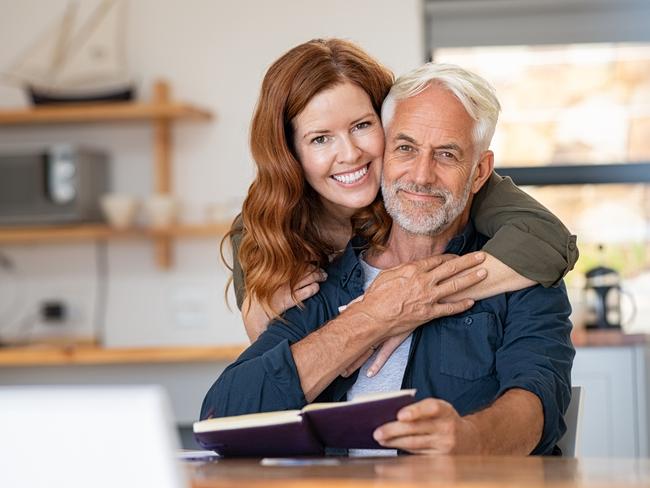 Portrait of happy mature couple at home looking at camera. Romantic wife embracing senior husband from behind while laughing together. Portrait of beautiful woman in love hugging  old man in perfect harmony. Retirement generic seniors