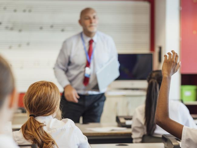 A rear-view shot of teenage music students in a classroom learning how to play the keyboard with their teacher at a school. The students are wearing school uniforms and the teacher is smartly dressed.