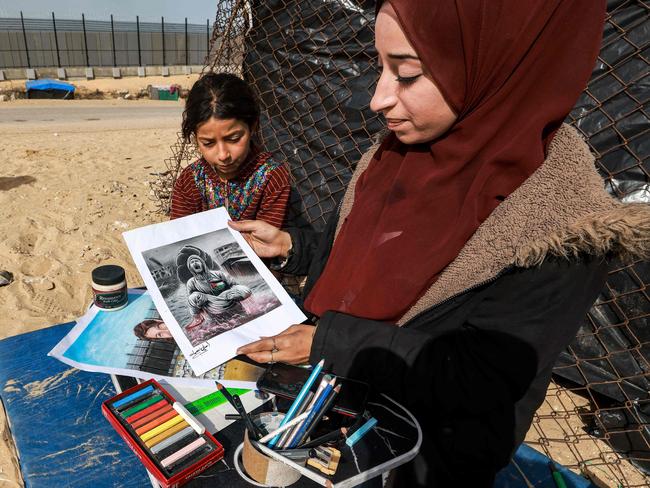 Palestinian artist Aseel Nasman (R) presents a painting as she sits alongside her younger sister Remas outside a tent at a camp for displaced Palestinians in Rafah. Picture: AFP