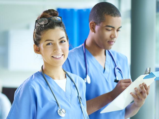 a young male and female nurse stand proudly on the ward and look to camera . In the background a patient is being attended by a nurse and their relative . Source: iStock