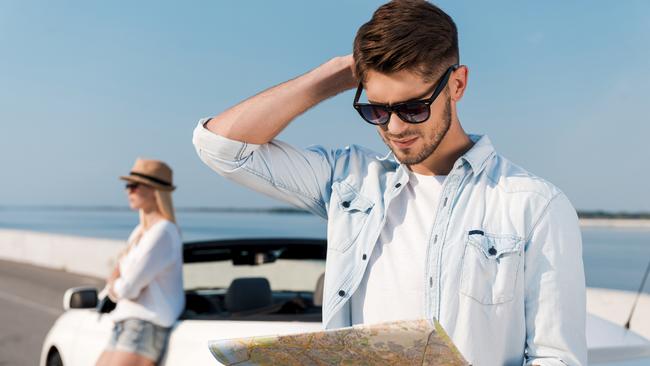 Trying to find their way. Frustrated young man examining map while his girlfriend leaning at their white convertible in the background. For Rod Chester column