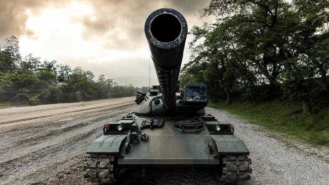 A wide-angle front view of a Japanese self-defense force military tank driving down a dirt road lined with trees with a dramatic sky.