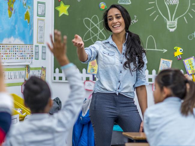 A teacher is leading a class of elementary school children. There are various posters on the wall, and drawings on the chalkboard. Students are putting up their hands to answer a question.