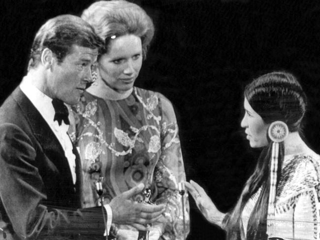 Sacheen Littlefeather (r), holds her hand out to Liv Ullmann and Roger Moore to refuse the Oscar for actor Marlon Brando who won the coveted movieland award for his part in 'The Godfather.' Miss Littlefeather said she has a letter from Brando explaining his refusal, saying that he refused the award because of the treatment by Americans of American indians. Oscars. Academy Awards.
