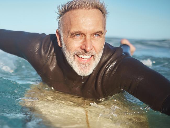 Beach, water and old man surfer swimming on a summer holiday vacation in retirement . Smile, ocean and senior surfing or body boarding enjoying a healthy exercise. Do the things you love to help your mental health.