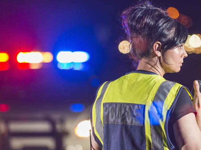 Rear view of a female police officer standing in the street at night, talking into her radio. Her patrol car is in the background with the emergency lights illuminated. She is wearing a yellow safety vest. Tablet generic