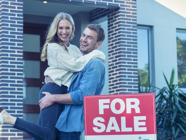 Couple in front of a new home. They are standing next to a for sale sign. They are both wearing casual clothes and embracing. They are smiling and he has a beard. He is lifting his wife off the ground with joy. The house is contemporary with a brick facade. The front door is also visible. Copy space