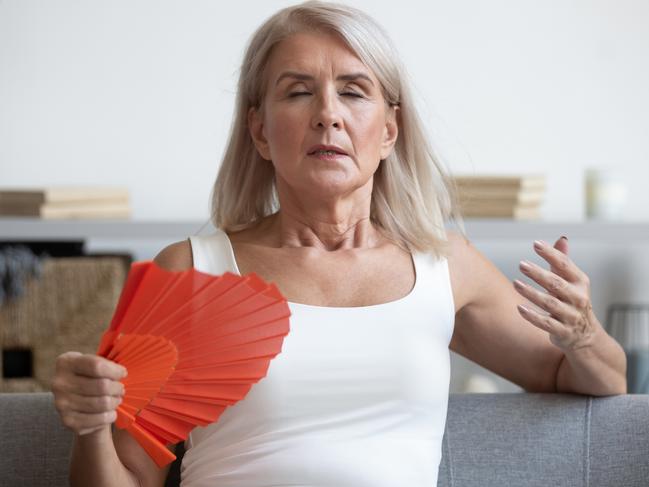 Tired mature woman suffering from heat at home, feeling uncomfortable, waving blue fan, sitting on couch, enjoying fresh air, sweaty older female cooling in hot summer weather, high temperature