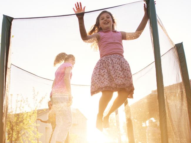 Three charming cute kids are jumping on the trampoline and the youngest girl is attempting to jump over the safety net during a beautiful sunny day.