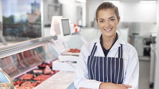 a young female butcher tends the meat counter in a butcher's shop. She is wearing a white coat and striped apron and is smiling to camera. Behind her the meat counter can be seen defocussed . butcher shop generic Savvy Shopper