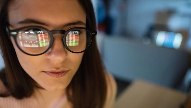 Close-up of a young woman looking at financial graphs on a laptop in a dark room with financial graphs reflected in her glasses.; investing generic money