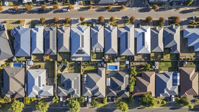 Aerial view directly above new outer suburban/semi-rural housing development with single-level housing between two streets with t-intersections and orange coloured street trees.  Mostly gray metal roofing, landscaped front and back yards, some solar panels, cars and motor vehicles parked in driveways and street, some street and parkland trees, water tanks in backyards, sheds and garages, swimming pools.  Mount Barker, South Australia; property investment housing money generic