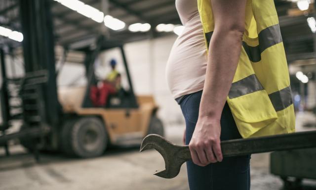 Pregnant woman working in construction factory. Man driving forklift in background