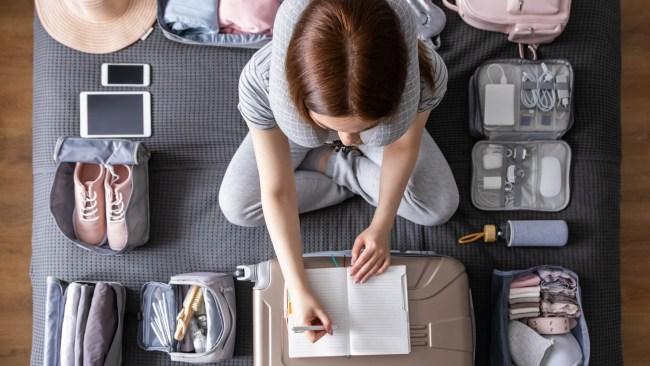 Smiling tourist woman packing suitcase to vacation writing paper list sitting on bed at home. Female getting ready to travel trip organization things storage in comfy cases bags use konmari method