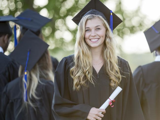 A multi-ethnic group of university students are outdoors at a graduation ceremony. They are wearing graduation robes and caps. They are walking away from the camera, and a Caucasian woman is turned to smile at the camera while holding her diploma.