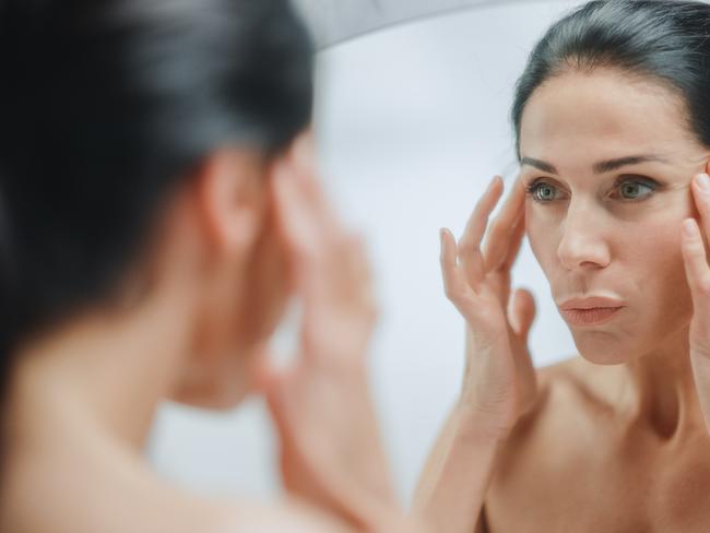 Caucasian Woman Looking in Mirror, Touching Beautiful Face, Soft Skin. Elderly Lady with Lush Black Hair. Female Beauty, Dignity, Grace. Sensual Wellness and Organic Skincare Products. Portrait Shot