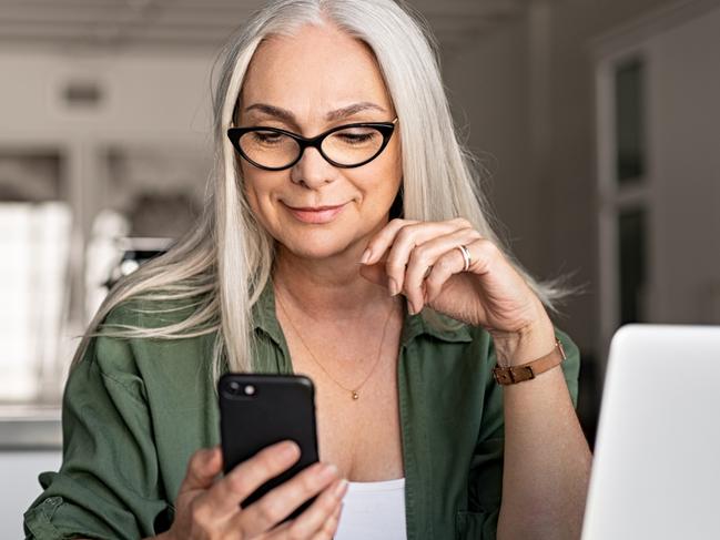 Happy senior woman using mobile phone while working at home with laptop. Smiling cool old woman with white hair wearing eyeglasses sitting on chair at table and messaging with smartphone. Beautiful stylish elderly lady browsing site on cellphone.  working from home istock