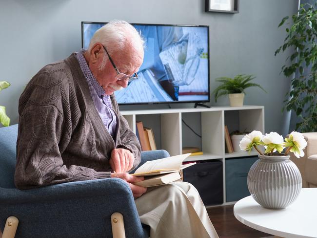 Elderly man in eyeglasses sitting in armchair and reading a book during his rest in the living room at home