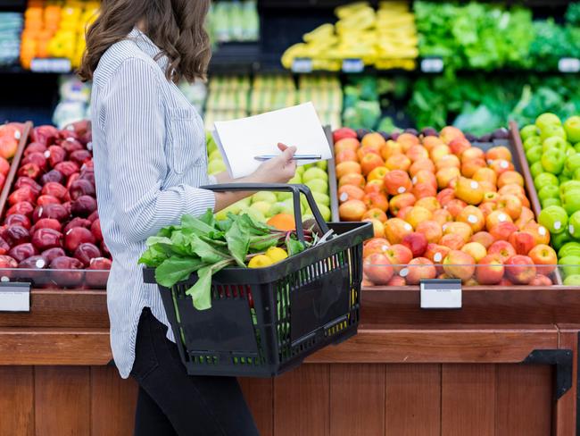 Young woman carries a shopping basket filled with fresh produce. She is shopping for fresh fruit and vegetables in a grocery store.