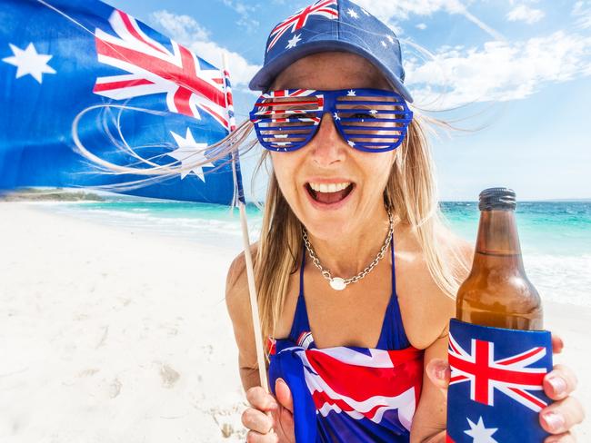 Australia Day national public holiday January 26.  Woman on beach wearing patrotic items of clothing bearing the Australian flag and she is also waving a small Australian flag and holding a drink in a cooler