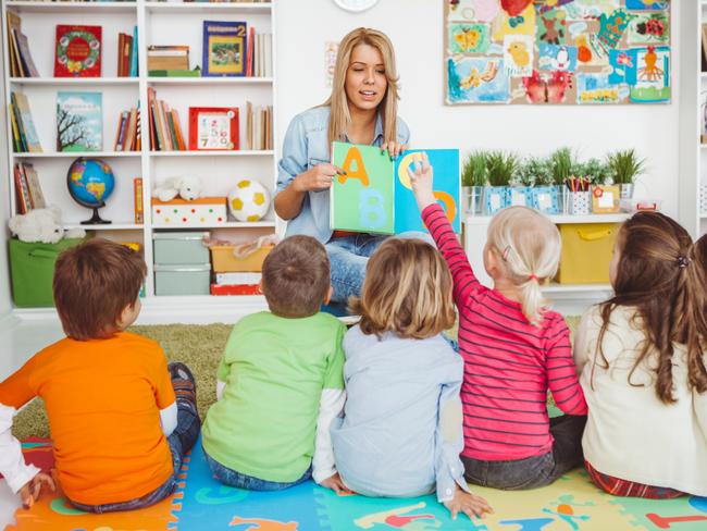 Teacher with a group of preschool children in a nursery. The children are sitting on the floor and listening  teacher. Learning letters. In the background we can see a shelf with some, toys, black board and books.