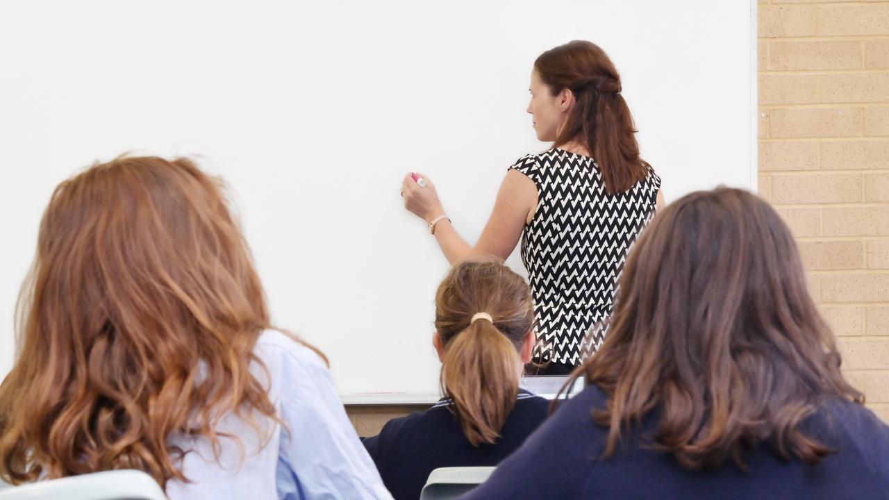 Rear view of primary school teacher writing on a clear whiteboard in classroom with students.