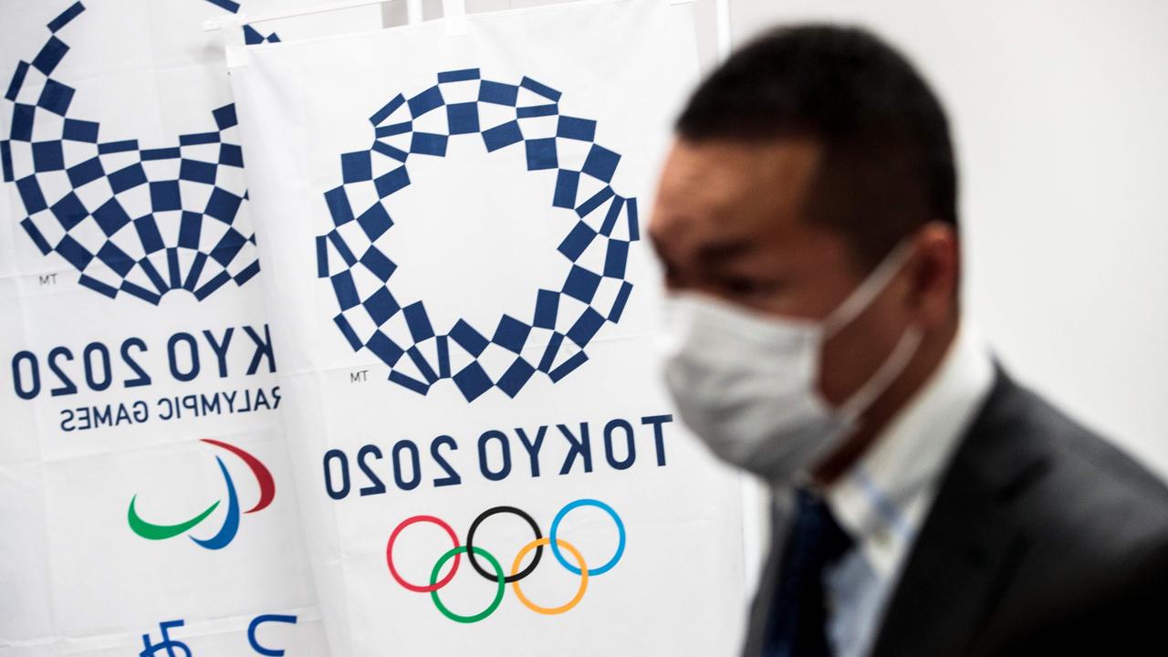 A reporter wearing a face mask stands next to the banners of the Tokyo Olympics.