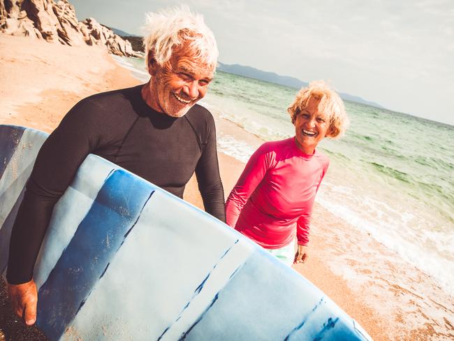 Very active senior man, walking down the beach while holding a surfboard under one armpit, and his female partner in the other hand.