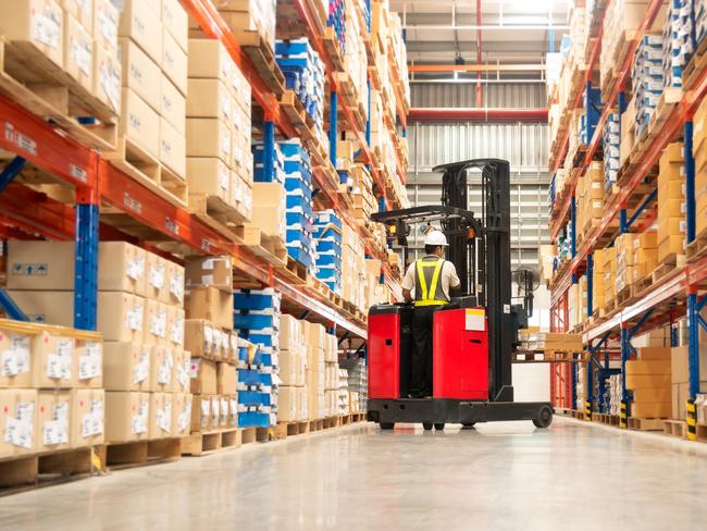 Worker in forklift-truck loading packed goods in huge distribution warehouse with high shelves.