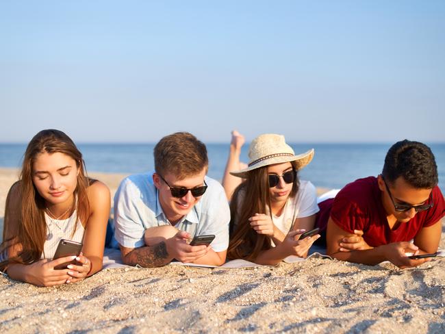 Group of millenials using smartphones laying together on beach towel near sea on summer sunset. Always connected generation communicate via internet. Young people addicted by mobile smart phones.