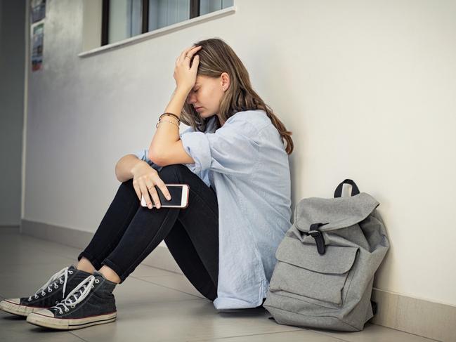 Upset and depressed girl holding smartphone sitting on college campus floor holding head. University sad student suffering from depression sitting on floor at high school. Lonely bullied teen in difficulty with copy space.