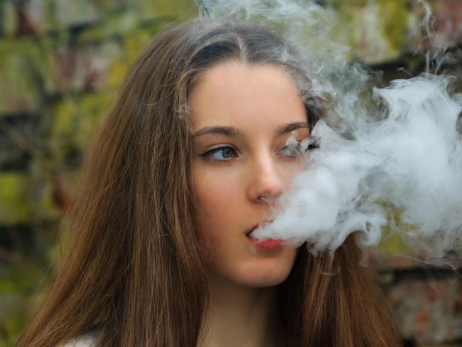 Vape teenager. Young pretty white girl in white cardigan smoking an electronic cigarette opposite destroyed brick wall on the street in the spring. Bad habit that is harmful to health.