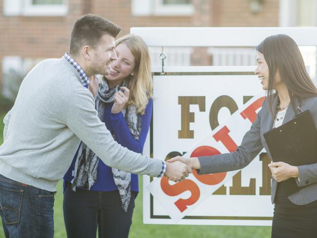 A Caucasian man and woman are buying a house together. They are standing outside the house with a sold sign and their real estate agent. The man and agent are shaking hands.