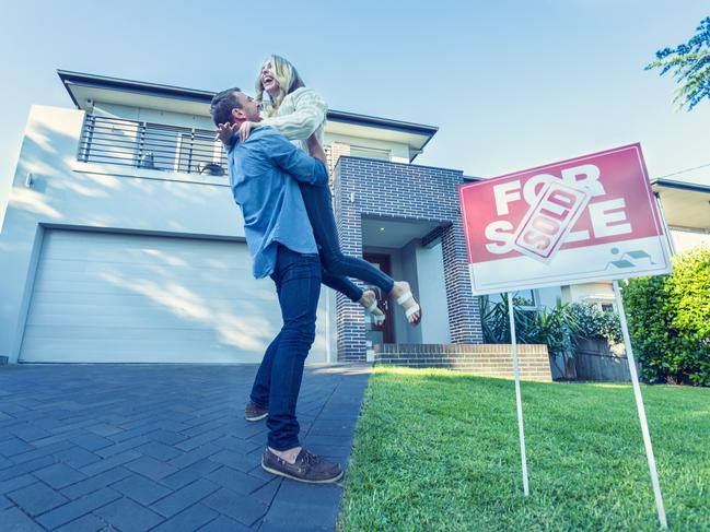 Couple standing in front of a new home. They are standing next to a for sale sign with a sold sticker. They are buying or selling this real estate. They are both wearing casual clothes and embracing. They are smiling and he has a beard. The house is new and contemporary with a brick facade. Copy space
