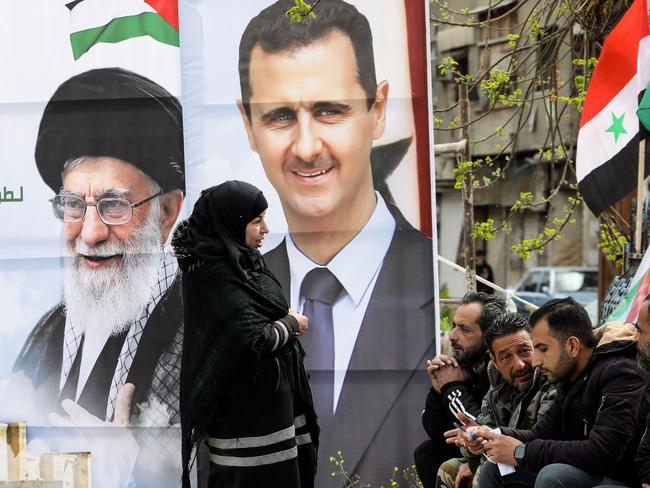 Portraits of Syria's President Bashar al-Assad (R) and Iran's supreme leader Ayatollah Ali Khamenei stand as Palestinians sit at the entrance of the Yarmuk camp for Palestinian refugees, south of Damascus, during a delivery of humanitarian aid provided by Iran as part of the Muslim holy fasting month of Ramadan on March 26, 2024. (Photo by LOUAI BESHARA / AFP)