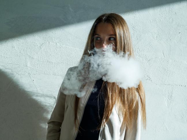 Vape teenager. Young cute girl in  casual clothes smokes an electronic cigarette near the wall outdoors in summer day. Bad habit that is harmful to health. Vaping activity.