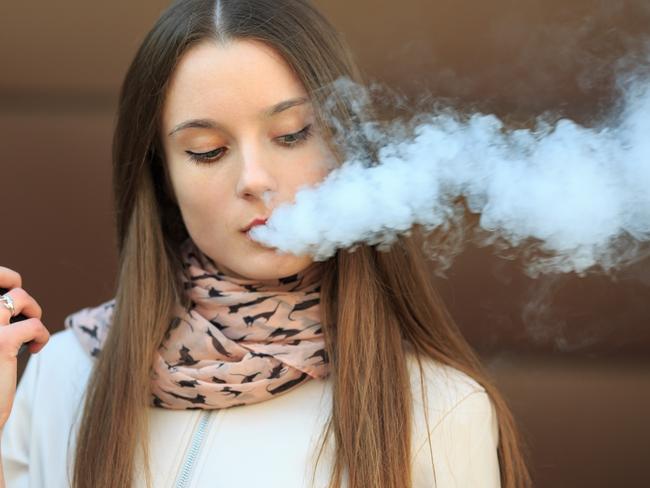 Vape teenager. Young pretty white girl in casual clothing smoking an electronic cigarette opposite modern brown background on the street in the spring. Bad habit. Vaping activity. Close up. i STOCK IMAGE