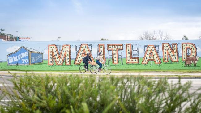 Couple riding their bicycles past the Welcome to Maitland Sign on Elgin St, Maitland.