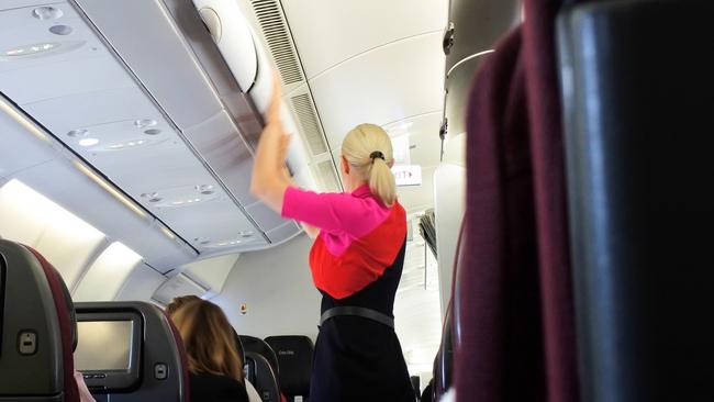 Brisbane - Oct 31 2022: Qantas airways female flight attendant closing the overhead luggage compartment lid for carry on baggage before take off.In Australia 1 flight attendant for every 36 passengers.Escape 16 June 2024Why I travelPhoto - iStock