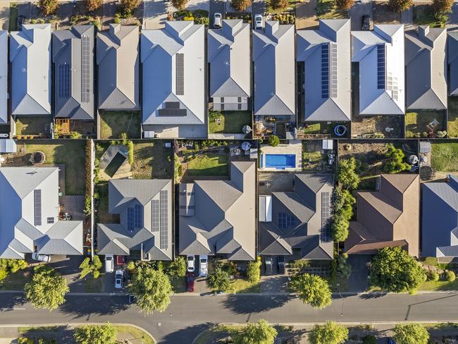 Aerial view directly above new outer suburban/semi-rural housing development with single-level housing between two streets with t-intersections and orange coloured street trees.  Mostly gray metal roofing, landscaped front and back yards, some solar panels, cars and motor vehicles parked in driveways and street, some street and parkland trees, water tanks in backyards, sheds and garages, swimming pools.  Mount Barker, South Australia; property investment housing money generic