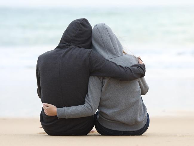 Rear view of a couple of pensive teenagers hugging and watching the sea sitting on the sand of the beach in a rainy day