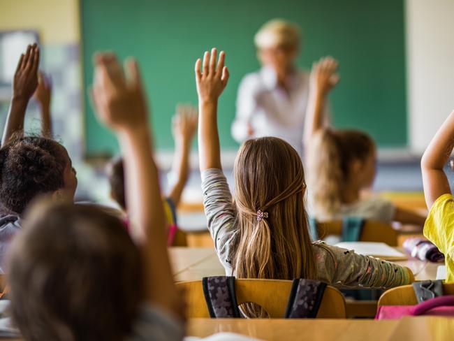 Rear view of large group of students raising their arms to answer the question on a class at elementary school.