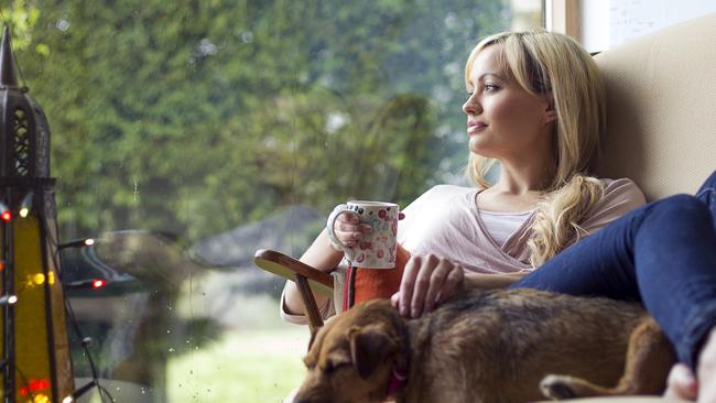 A happy blonde woman lies down on her sofa at home with a hot drink in her hand while looking out of her window. Her dog is lying next to her asleep. Ornaments and lights can be seen in the room.