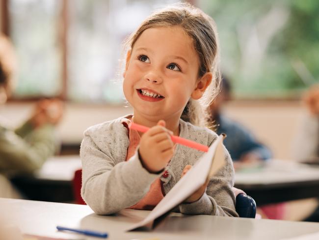 Schools of excellence - Excited little girl learns to draw with a colour pencil in an art class. Happy elementary school kid talks to her teacher while holding a colour pen and a drawing book. Creativity as a part of primary education.