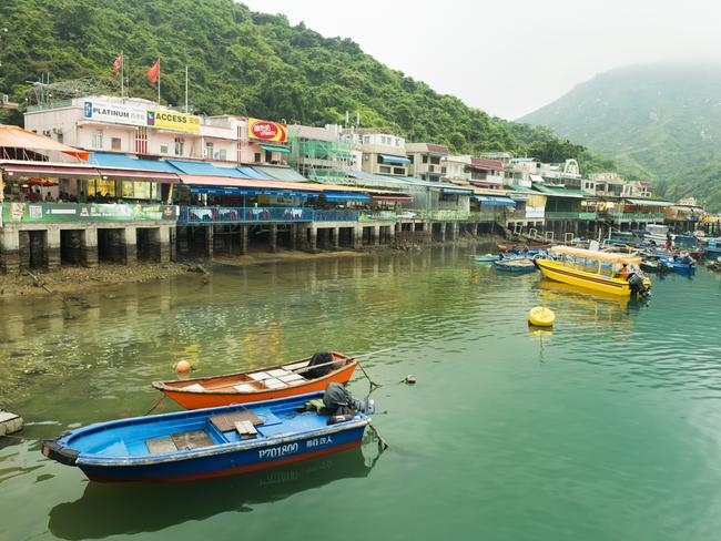 This is a horizontal, color photograph of the scenic harbor and village in Sok Kwu Wan Village on Lamma Island in Hong Kong. Colorful boats float on the water and green mountains disappear into the fog in the background.